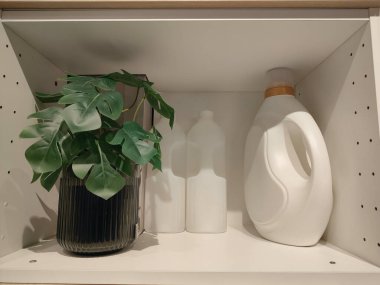 Rafta beyaz bir çamaşır sepeti saksı bitkisi ve bir şişe deterjanın yanında duruyor.