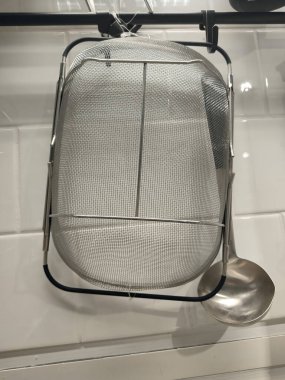 A silver mesh strainer is hanging from a hook on a white wall. A spoon is sitting on the counter next to the strainer clipart