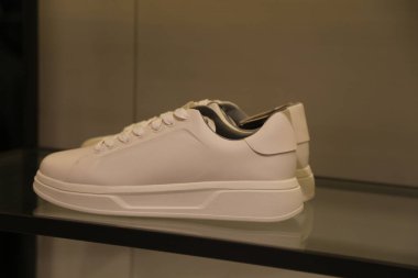 Bir rafta bir çift beyaz spor ayakkabı sergilenir. Ayakkabılar temiz ve bakımlı, siyah tabanlı ve üst kısmı beyaz. Spor ayakkabıları gündelik kıyafetler için tasarlanmış.