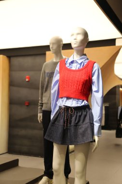 Bir mağazada bir erkek ve bir kadın yan yana duruyorlar. Adam gri bir kazak giyiyor ve kadın da mavi gömlek ve kırmızı bir yelek giyiyor.