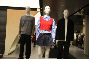 Üç manken bir mağazada duruyor, farklı kıyafetler giyiyorlar. Mankenler bir süveter, bir etek ve bir ceket de dahil olmak üzere çeşitli giysiler giyiyorlar. Mağaza iyi aydınlatılmış.