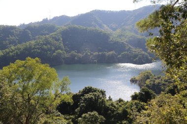 Ağaçlar ve dağlarla çevrili güzel bir göl. Göl sakin ve huzurlu, güneş sudan yansıyor. Ağaçlar yemyeşil bir zemin oluşturuyor, dingin bir zemin oluşturuyor.