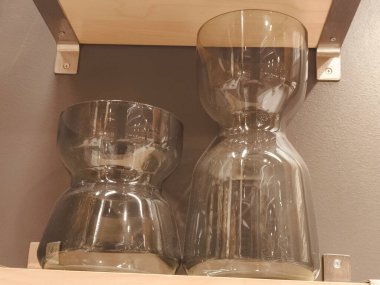 Bir rafta iki vazo, biri diğerinden daha uzun. Vazolar camdan yapılmış ve bir rafta duruyor.