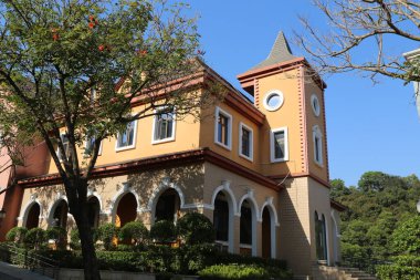 Saat kulesi ve geniş penceresi olan büyük bir bina. Bina turuncu ve beyaz. Binanın arka planında bir sürü pencere ve bir sürü ağaç var.