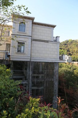 A tall building with a balcony and a large window. The building is surrounded by trees and bushes