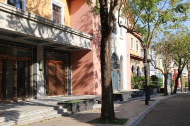 Bir sıra bina ve ortasında bir ağaç olan bir cadde. Sokak dükkanlarla dolu ve kaldırımı var.
