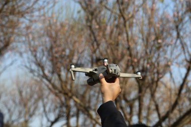 Bir insan elinde bir dron tutuyor. Drone gri ve siyah ve kırmızı bir pervanesi var. Sahne arka planda ağaçlar olan bir parkta geçiyor.