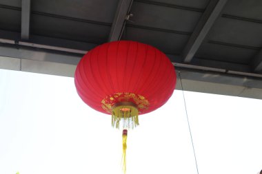 Sarı süslemeli kırmızı bir fener bir binadan sarkıyor. Fenerin ışığı yanıyor ve çevresine sıcak bir parıltı yayıyor. Fenerin tasarımı ve rengi şenlikli ve davetkar bir atmosfer yaratır.