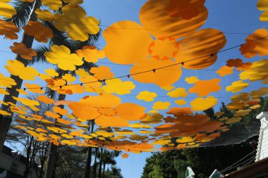 Bir caddenin üzerinde büyük bir portakal çiçeği örtüsü asılı. Çiçekler havada süzülüyormuş gibi görünecek şekilde düzenlenmiş. Sahnenin kaprisli ve neşeli bir havası var.