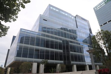 Önünde yeşil ağaç olan büyük camdan bir bina. Bina çok yüksek ve birçok penceresi var, modern ve şık bir görünüme sahip. Binanın önündeki ağaç doğaya bir dokunuş ekliyor.