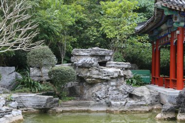 Küçük gölü ve bankı olan bir kayalık bahçe. Gölet kayalar ve bitkilerle çevrili ve yakınlarda bir bank var. Sahnenin huzurlu ve huzurlu bir atmosferi var.