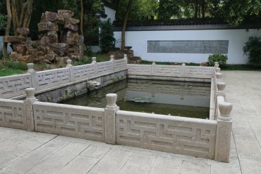 Avludaki beyaz taş çitin içinde küçük balıklar ve koiler yüzüyor. Basit bir desen ve gri tonda Çin tarzı korkuluklar var..