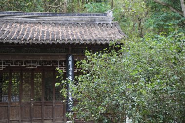 Ormanda Çin tarzı bir bina, çatısında koyu ahşap ve siyah fayanslar var. Bir tarafındaki duvarlar oyulmuş pencereler ve ona açılan açık bir kapı..