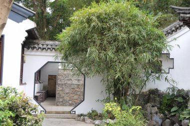 Yeşil bitkiler ve bambu ağaçlarıyla çevrili açık kapısı olan beyaz, Çin tarzı bir avlu. Evin duvarlarının üstünde siyah fayanslar ve altında beyaz taş duvarlar var..