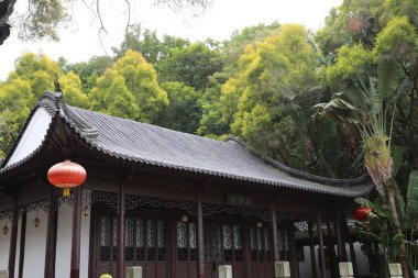 Parkta yeşil ağaçlar ve bitkilerle çevrili Çin tarzı bir bina. Çatı koyu gri fayanslarla kaplı ve üzerinde turuncu bir fener asılı..