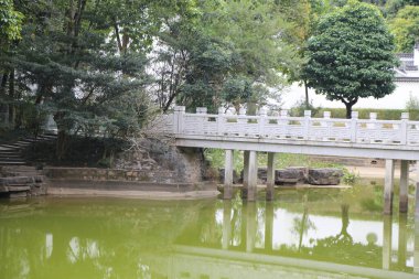 Beyaz bir köprü yeşil göletin üzerinde kadim bir Çin bahçesinde kavis çiziyor, ona doğru giden taş adımlar ve yolun her iki tarafında da ağaçlar var..