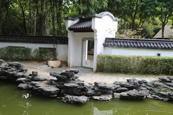 Beyaz kapısı ve gölete giden taş yolu olan taş bir duvar. Kapı açık ve patika çalılarla kaplı.