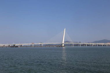 Bir köprü, üzerinde açık mavi bir gökyüzü olan bir su kütlesini kaplar. Köprü, uzaktan görülebilen büyük, beyaz bir yapıdır.