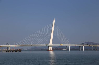 Bir köprü, üzerinde açık mavi bir gökyüzü olan bir su kütlesini kaplar. Köprü beyaz ve uzun, ince bir tasarımı var. Köprünün altındaki su sakin ve huzurlu, görünür bir tekne ya da başka bir aktivite yok.