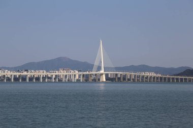 Bir köprü geniş bir su kütlesi boyunca uzanır ve arka planda bir şehir vardır. Köprü beyazdır ve suyun tüm genişliğini kaplar.