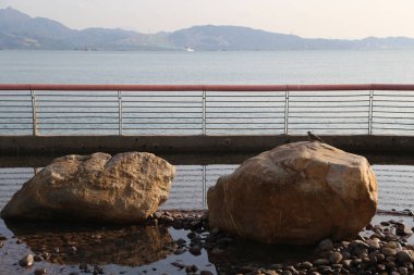 Arka planda parmaklık olan iki büyük kaya bir gölette oturuyor. Su sakin ve gökyüzü berrak.
