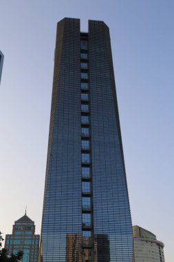Çok yüksek bir bina ve arka planda açık mavi bir gökyüzü var. Bina o kadar yüksektir ki çevreyi kule gibi sarar. Pencereler büyük ve sayısız.