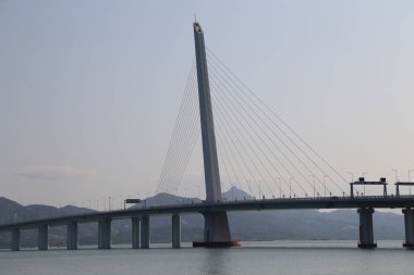 Bir köprü, üzerinde açık mavi bir gökyüzü olan bir su kütlesini kaplar. Köprü uzundur ve üzerinde birkaç araba bulunan geniş bir mesafeyi kapsar.