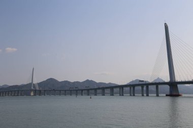 Bir köprü, üzerinde açık mavi bir gökyüzü olan geniş bir su kütlesini kapsar. Köprü dağlarla çevrili, resim gibi bir manzara yaratıyor.