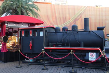 Kırmızı süslemeli büyük, siyah bir tren bir platforma park edilmiştir. Trenin üzerinde kırmızı bir gölgelik var, elementlere karşı gölge ve koruma sağlıyor. Trendeki bir işaret sigara içilmemesi konusunda uyardı.