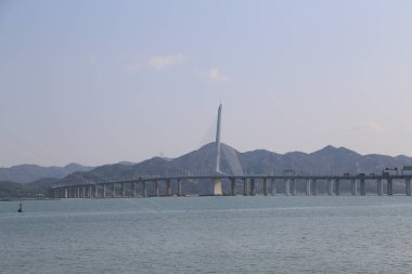 Bir köprü geniş bir su kütlesi boyunca uzanır, arka planda dağlar vardır. Köprü, suyun üzerinde asılı duran uzun ve ince bir yapıdır. Gökyüzü açık ve su sakin.