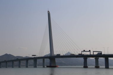 Bir köprü, üzerinde açık mavi bir gökyüzü olan bir su kütlesini kaplar. Köprü dağlar ve ormanlarla çevrili.