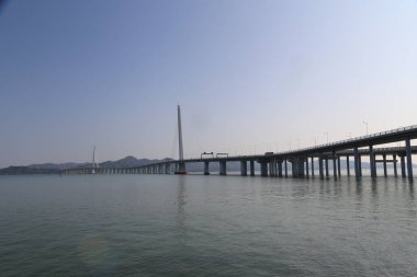 Bir köprü, üzerinde açık mavi bir gökyüzü olan bir su kütlesini kaplar. Köprü uzundur ve geniş bir mesafe kateder.