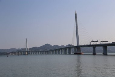 Uzun bir köprü, üzerinde açık mavi bir gökyüzü olan bir su kütlesi boyunca uzanır. Köprü dağlarla çevrili, güzel bir manzara yaratıyor.