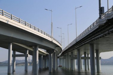 Bir köprü, üzerinde açık mavi bir gökyüzü olan bir su kütlesini kaplar. Köprü geniş, çok genişlikli bir yapıdır ve birkaç ışık mesafesi görülebilir.