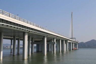 Bir köprü, arka planında uzun bir kule olan bir su kütlesini kapsar. Köprü beton ve çelikten yapılmıştır ve çok uzun bir yapıdır. Su sakin ve hareketsiz, köprüyü yansıtıyor.