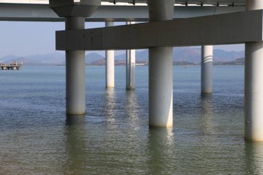 Uzun bir köprü bir su kütlesi boyunca uzanıyor, yansıması suda görülebiliyor. Köprü beyaz malzemelerden yapılmıştır ve sakin, mavi bir okyanusla çevrilidir.