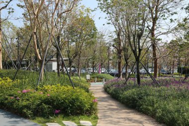 İçinde patika ve ağaçlar olan bir park. Yol çiçekler ve çalılarla doludur. Arka planda park etmiş birkaç araba var.