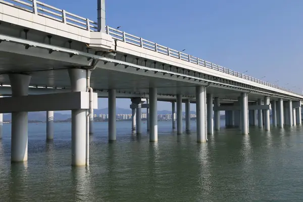 Uzun bir köprü, köprünün yapısını yansıtan suyla birlikte geniş bir su kütlesi boyunca uzanır. Köprü metalden yapılmıştır ve çok yüksektir, aşağıdaki suyun üzerinde yüksektir. Su sakin.