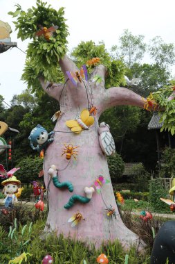 Renkli bahçede, arka planda yeşil ağaçlar ve çiçeklerle çevrili, üzerinde çeşitli çizgi film karakterleri asılı, aşırı büyük pembe bir ağaç gövdesi bulunuyor..