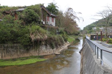 Kırmızı çatılı bir ev nehrin yanındaki tepede yer alır. Ev ağaçlar ve çalılarla çevrili.
