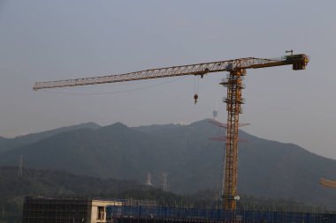 Büyük bir vinç bir dağ sırasının arka planındadır. Vinç sarı ve bir şey kaldırmak için kullanılıyor. Gökyüzü açık ve arka planda dağlar var.