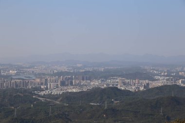 Uzakta, Shenzhen şehrinin manzarasını yüksek bir yükseklikten görebilirsiniz, aşağıdaki yeşil dağlar ve şehir binaları ile. Gökyüzü bulutsuz mavidir ve üzerinde bazı sisli hava katmanları var gibi görünüyor..