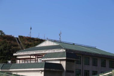 Çatıda güneş panelleri olan yeşil tepeleri ve açık mavi gökyüzünü gören bir bina. Sahne bu iki özelliği de vurgulayan bir açıdan çekiliyor. Önünde iki küçük televizyon kulesi var..