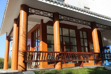 İki büyük pencerenin önünde ve beyaz tuğlalı bir evin en üst katındaki balkonda birbiri ardına basit çizgiler olan turuncu-kahverengi bir ahşap tahıl sütunu var..