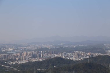 Şehrin uzak bir manzarası, önlerinde dağlar ve üzerlerinde binalar var. Gökyüzü bulutsuz ve sissiz açık mavi. Arkasında bir dağ sırası daha var..
