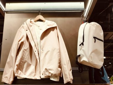 Beyaz bir ceket ve beyaz bir sırt çantası askıda asılı. Ceket uzun kollu, fermuarlı kapüşonlu bir ceket. Sırt çantası beyaz ve siyah bir kayışı var. Sahne bir mağazada geçiyor.