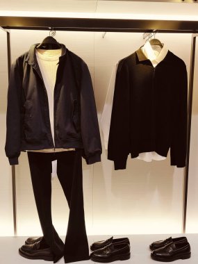 Siyah ceket, beyaz gömlek ve duvarın önündeki ayakkabılı bir askıda siyah pantolon. Ceket, smokin giyen bir mankenle minimalist bir odada askıda asılı duruyor.