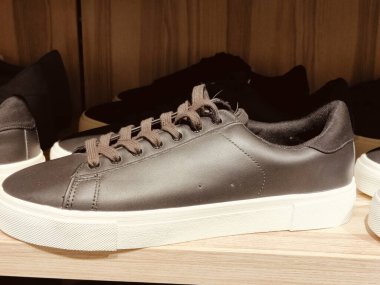 Koyu gri, üst, beyaz tabanlı ve kahverengi bağcıklı spor ayakkabılar bir mağaza ortamında sergilenmektedir. Ayakkabıların alçak ve ince tabanları var. Onlara zarif bir görünüm veriyor..