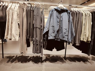 Stone Island mağazasındaki siyah metal askılarda sergilenen kapüşonlu, tişörtlü, kot pantolonlu ve şortlu kadınlar. İnce, koyu grimsi bir parıltısı var.