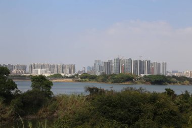 Nehrin karşısındaki şehrin ufuk çizgisi Çin sanayi parkındaki binalara, bitki örtüsüne ve gölün etrafındaki ağaçlara bakıyor. Arka planda uzun konut kuleleri, gün ışığı görülebiliyor.